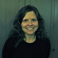 Maria smiles into the camera. She has brown curly hair to past her shoulders. Behind her is green paneling.