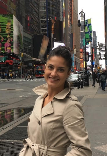 Ashley poses on a city street. She is wearing a khaki trench coat and her brown hair is pulled back.