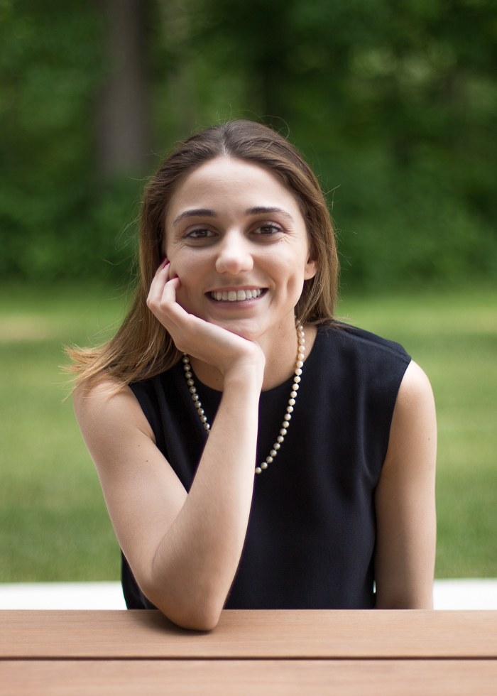 Laura rests her chin on her hand and smiles at the camera. She is outside, wearing a black sleeveless top and a chain necklace. She has light brown hair parted in the center.